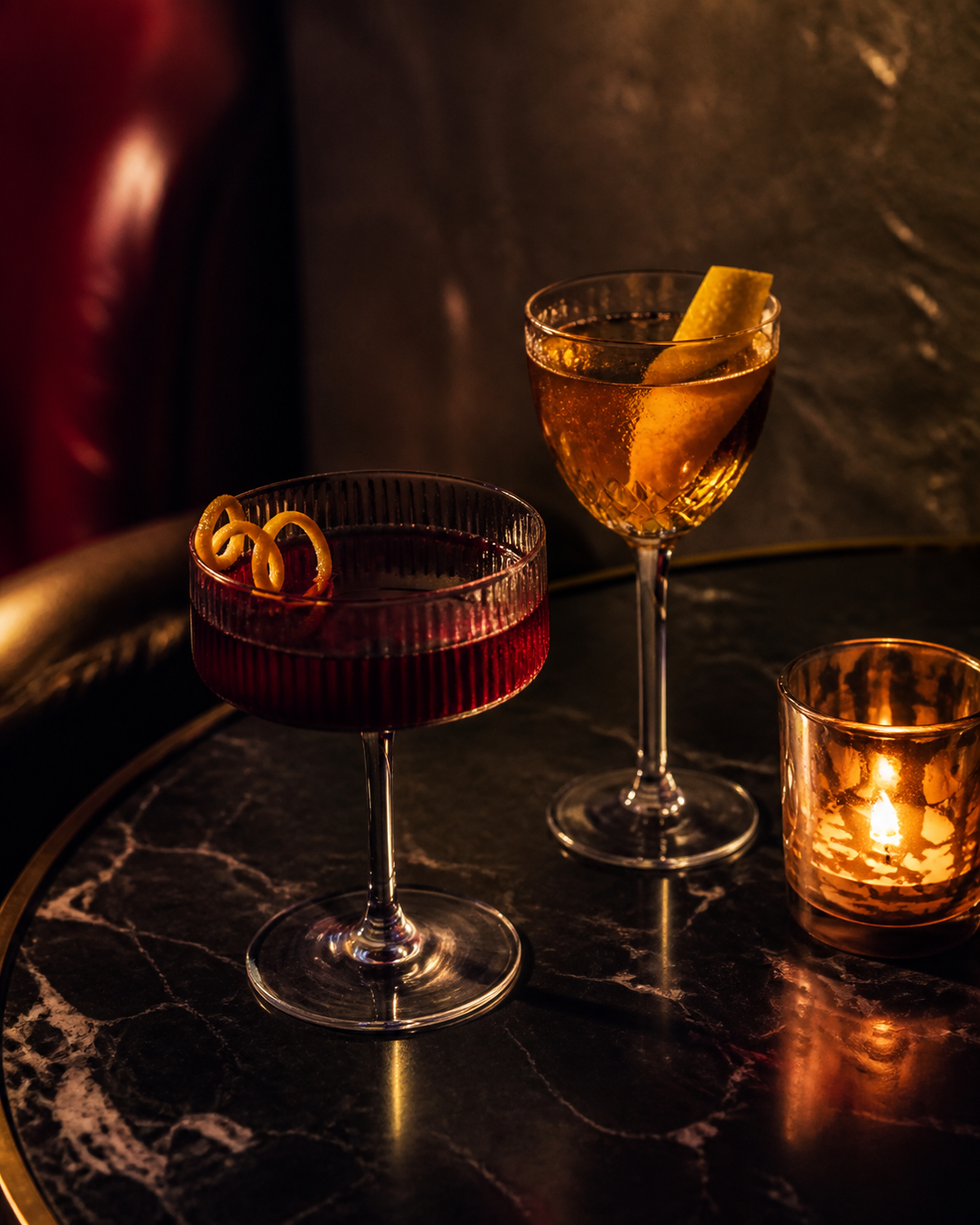 Two richly coloured cocktails and a candle on a dark marble table